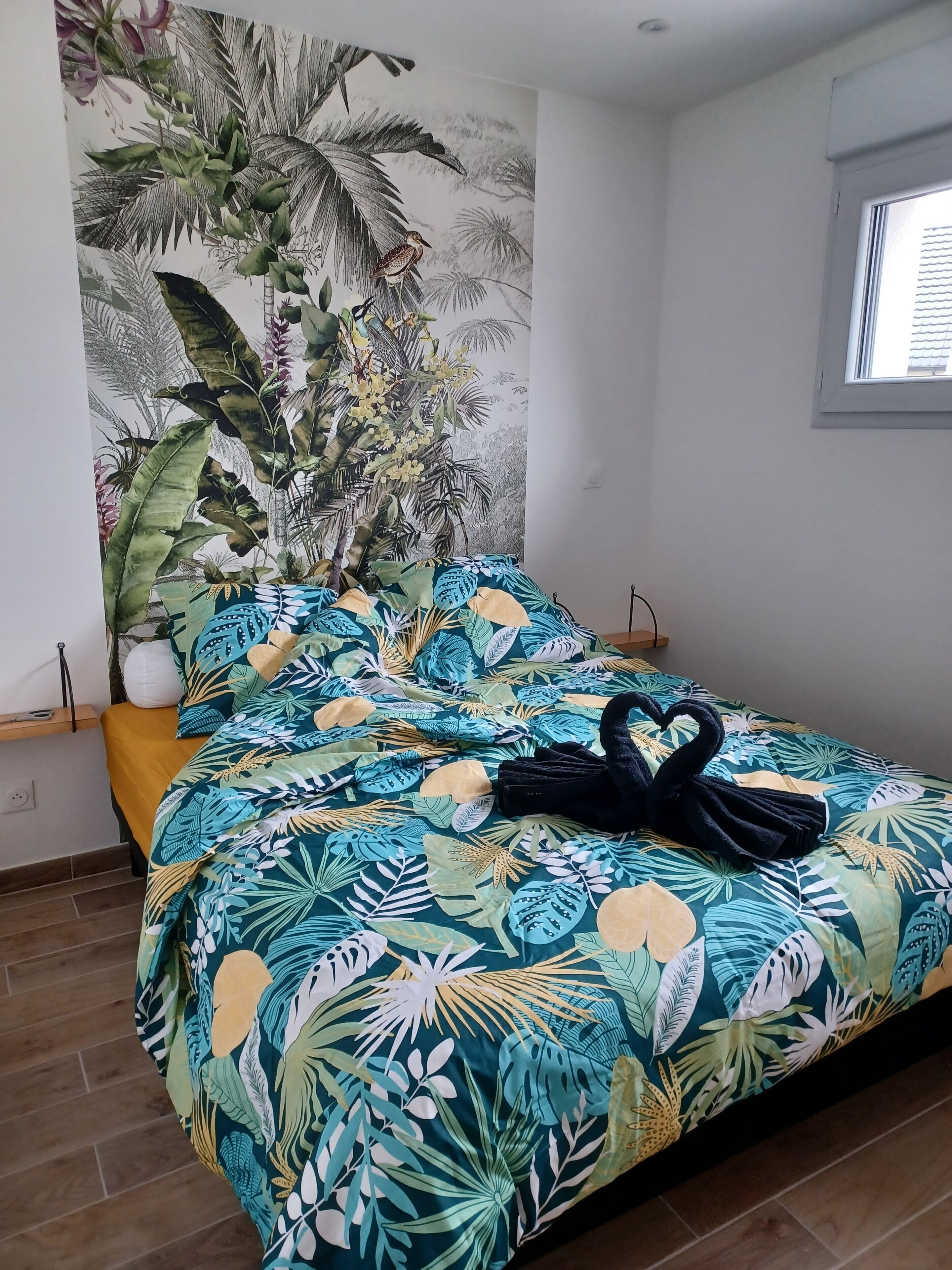 Un lit recouvert d’une couvre-lit ornée de motifs tropicaux, une serviette noire et un vase blanc posés sur la table de chevet.