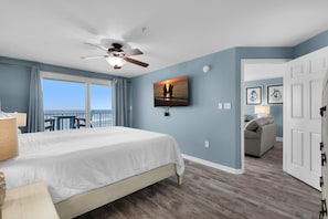 A bedroom with a bed, a ceiling fan, a TV mounted on the wall, and a view of the ocean through large windows.