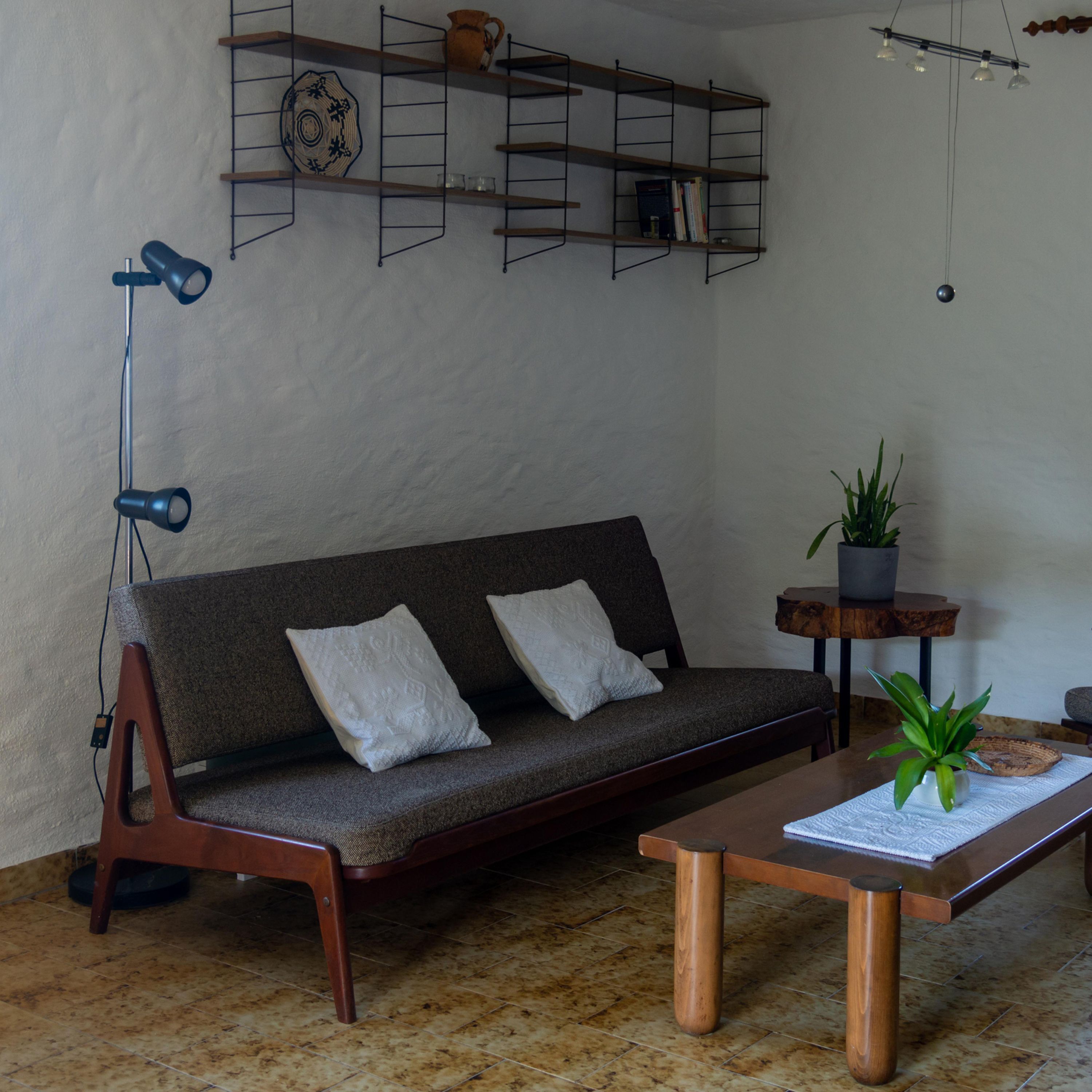 A modern living room with a sofa, a coffee table, a floor lamp, a small table with a plant, and a wall-mounted shelf with books and decorative items.