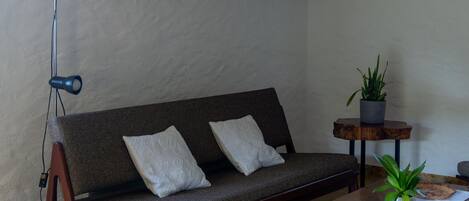 A modern living room with a sofa, a coffee table, a floor lamp, a small table with a plant, and a wall-mounted shelf with books and decorative items.