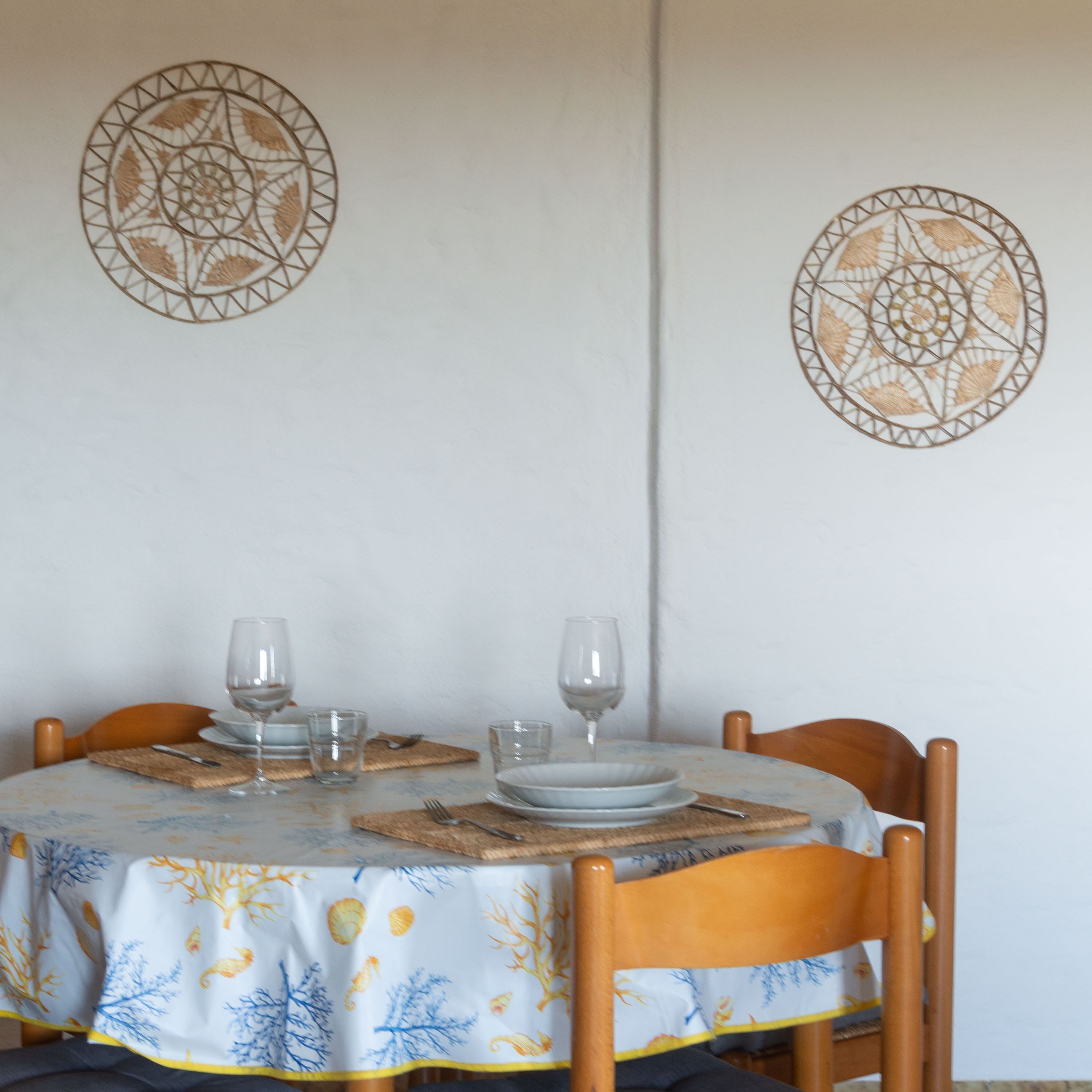 Uma mesa de jantar com toalha de mesa florida, quatro cadeiras e dois objetos decorativos na parede.