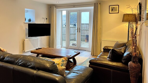Un salon avec un canapé en cuir, une table basse en bois, un téléviseur fixé au mur et une porte-fenêtre coulissante donnant sur un espace extérieur.