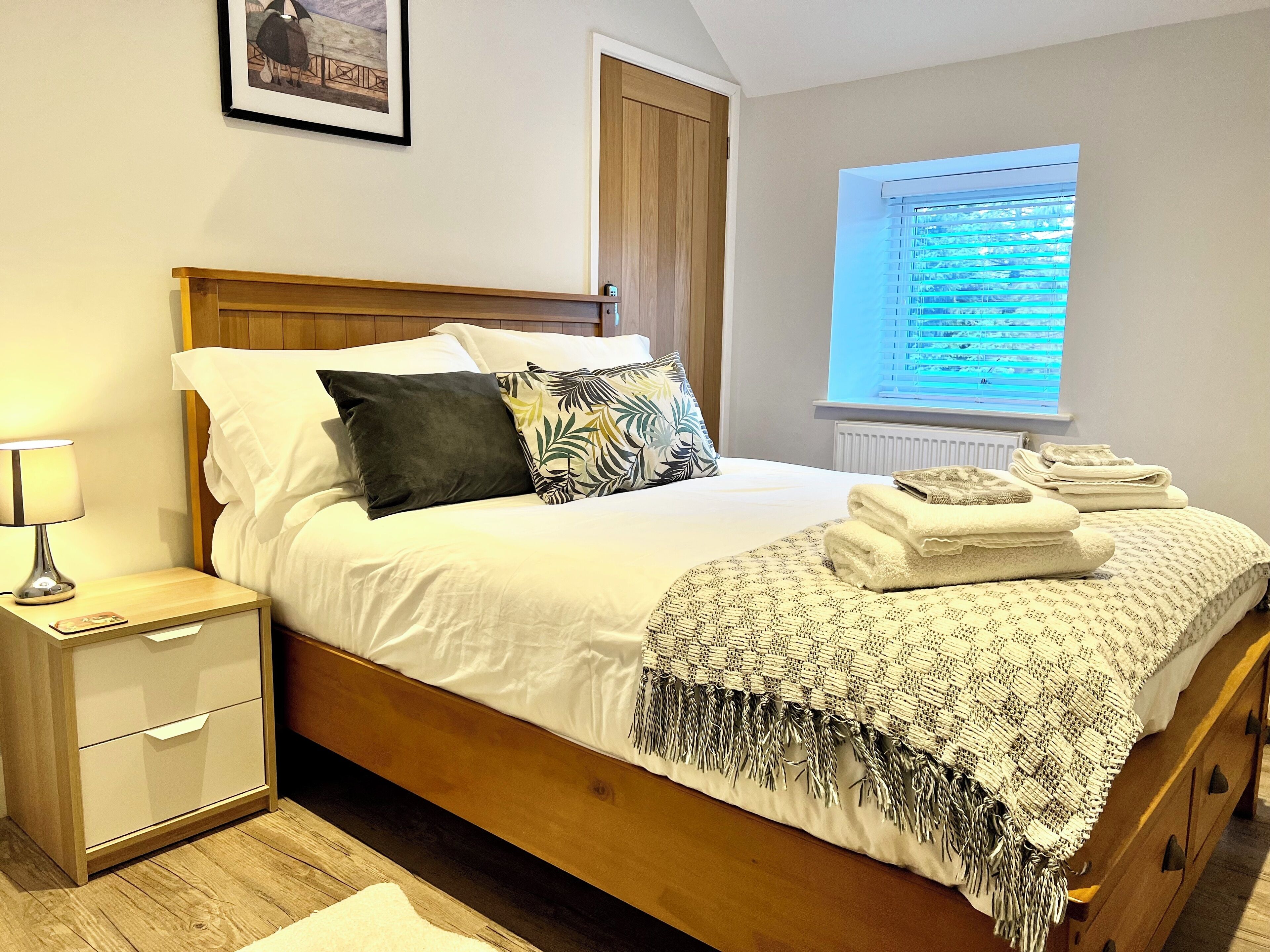 A neatly made bed with a white comforter, a dark pillow, and a tropical leaf-patterned pillow. There is a bedside table with a lamp and a folded towel. A window with blinds is visible, and a framed picture hangs on the wall.