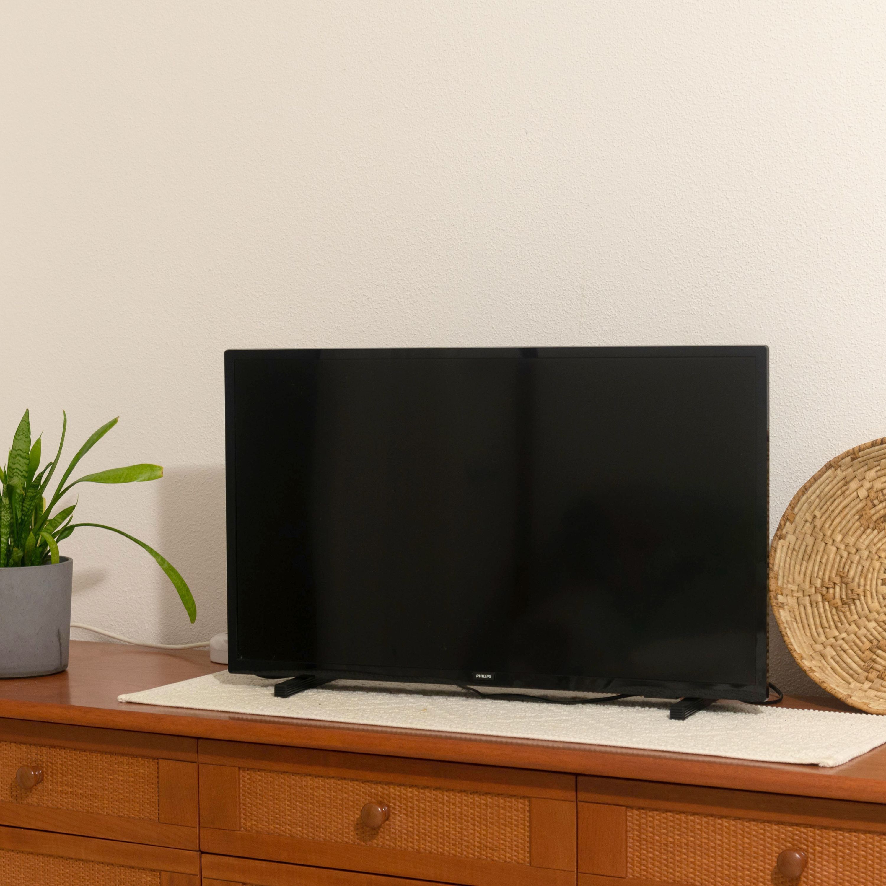 Un téléviseur à écran plat posé sur une commode en bois surmontée d’un tapis blanc, une plante en pot et un panier tressé.