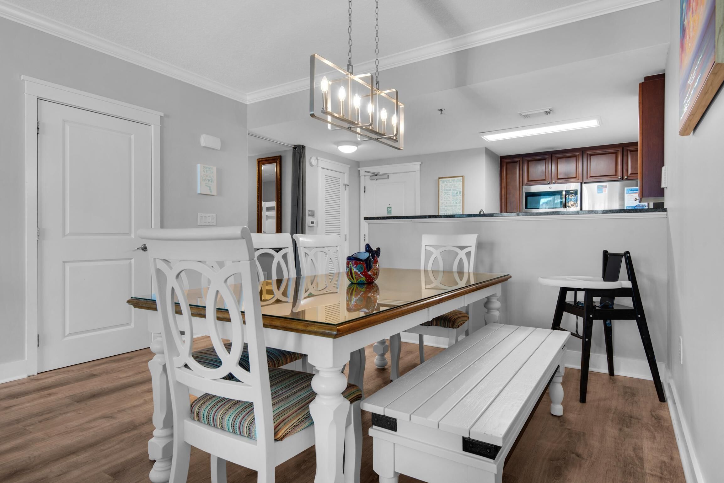 A dining area with a wooden table, white chairs, and a bench. There is a kitchen in the background with wooden cabinets and a microwave.