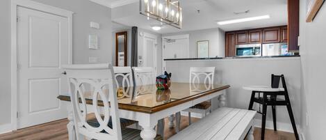 A dining area with a wooden table, white chairs, and a bench. There is a kitchen in the background with wooden cabinets and a microwave.