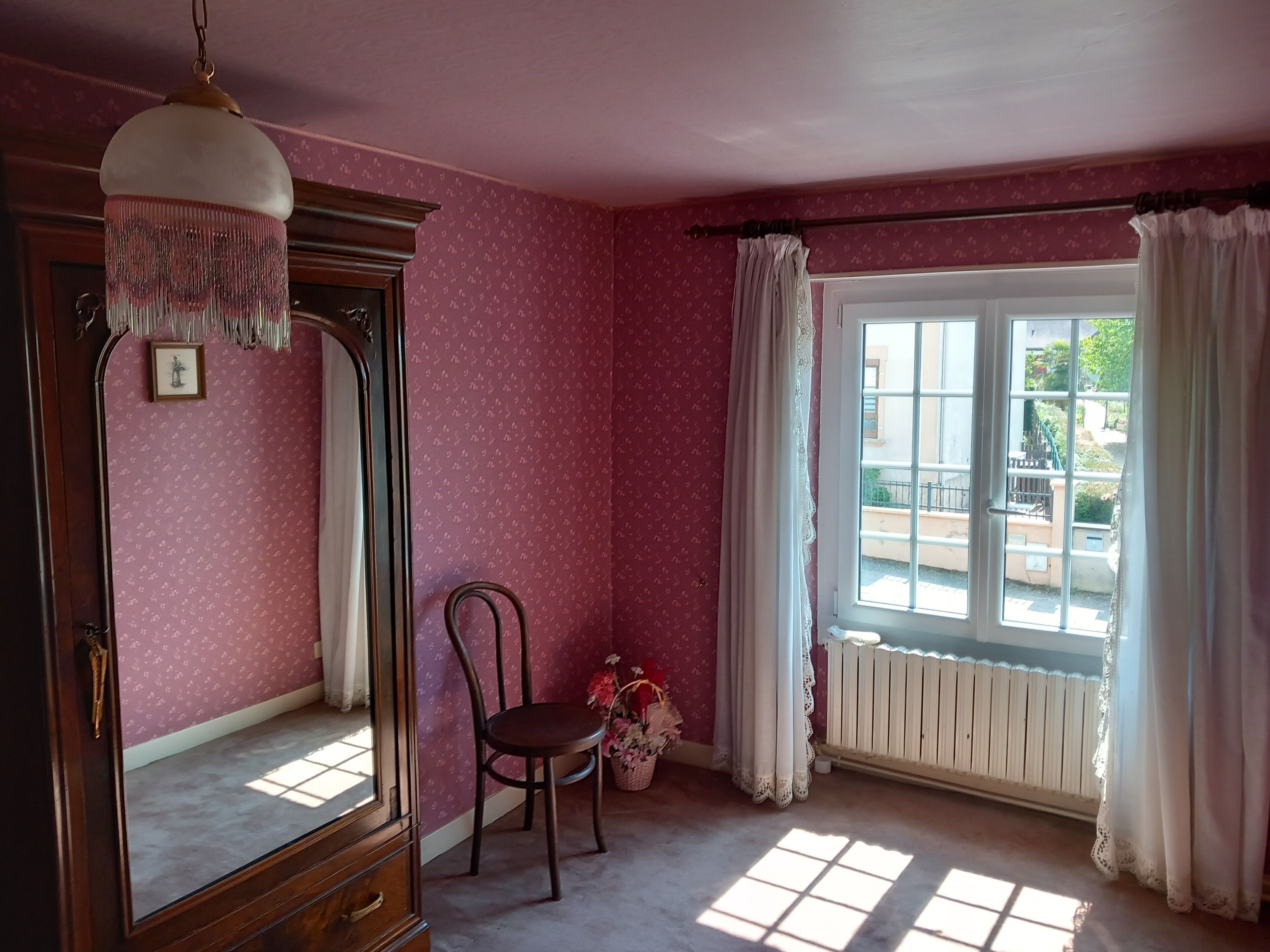 A room with pink wallpaper, a wooden dresser, a chair, and a window with curtains.