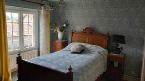 Een slaapkamer met een houten bed, een nachtkastje met een lamp, een vaas met bloemen en een raam met gordijnen.
