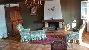 Een gezellige woonkamer met een open haard, fauteuils met bloemenmotief, een houten kast en een wanddecoratie van een pauw.