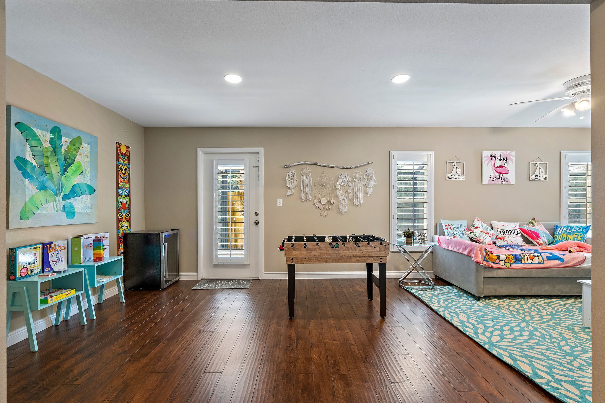 A living room with a foosball table, a sofa, and a coffee table. There are two windows with blinds, a door, and a wall with decorative items.