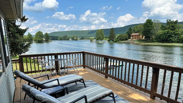 Terraza de madera con sillones y una mesa con vista a un lago, rodeada de follaje y colinas.