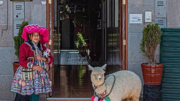 A person in traditional attire standing next to a llama outside a building with a large wooden archway and a sign with a 'H'. - Hotel Willcamayu  (Pisac)