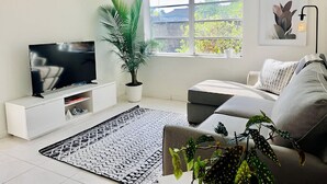 Un soggiorno moderno con una televisione a schermo piatto su un mobile bianco, un divano grigio, un tappeto decorato, una pianta di palma in vaso e una finestra con vista su piante.