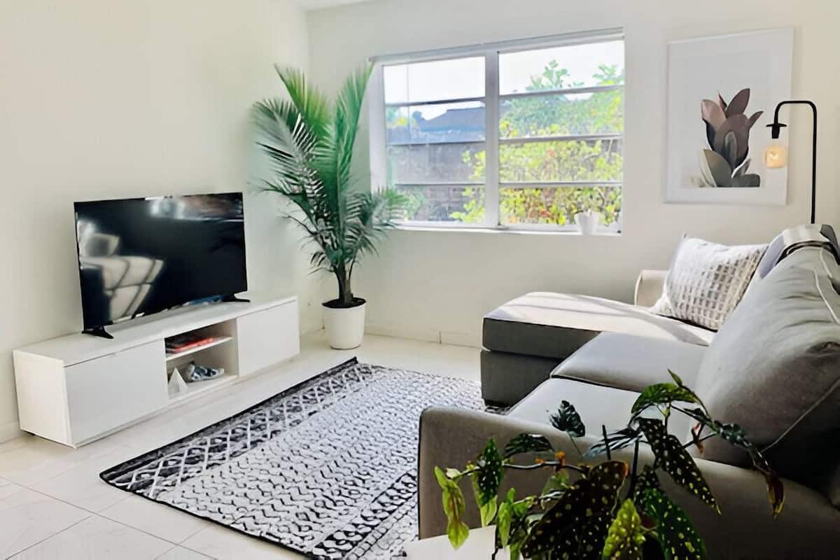 Un salon moderne avec un téléviseur à écran plat posé sur un meuble blanc, un canapé gris, un tapis à motifs, un palmier en pot et une fenêtre donnant sur un paysage verdoyant.