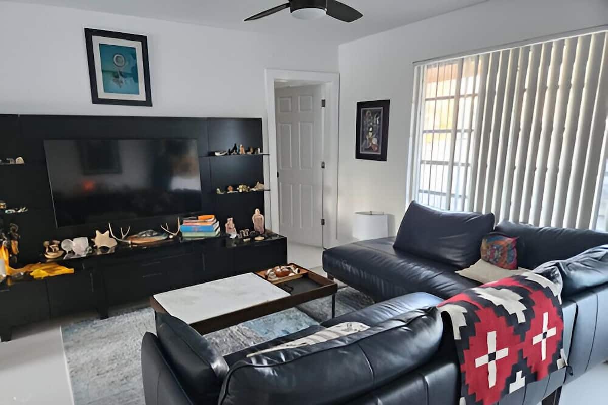 Un salon moderne avec un canapé en cuir noir, une table basse, un meuble de télévision noir agrémenté d’objets décoratifs et un grand téléviseur à écran plat.