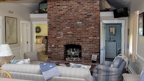 A cozy living room with a brick fireplace, two sofas, and a television mounted on the wall. - Cozy Ocean Side Cottage with screened in Sun room! 5 minutes from Private beach (Georgetown)