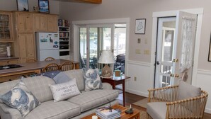 A living room with a sofa, coffee table, and a wicker chair. There is a kitchen area with wooden cabinets and a refrigerator. A sliding glass door leads to an outdoor area. - Cozy Ocean Side Cottage with screened in Sun room! 5 minutes from Private beach (Georgetown)