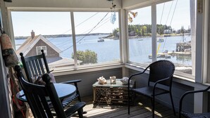 A wooden deck with two chairs and a table, overlooking a body of water with boats and houses in the distance. - Cozy Ocean Side Cottage with screened in Sun room! 5 minutes from Private beach (Georgetown)