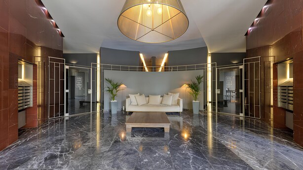 A spacious lobby with a marble floor, a central seating area with a sofa and coffee table, and a large, modern chandelier.