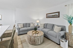 Uma sala de estar moderna com um sofá modular cinzento, uma mesa de centro redonda e uma área de refeições com uma mesa branca.