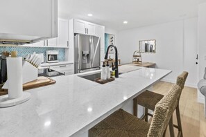 Een moderne keuken met witte kastjes, een roestvrijstalen koelkast, een marmeren werkblad en een geweven stoel.