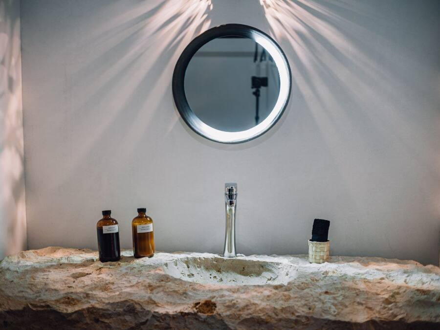 Un tocador de baño con un espejo redondo, un grifo y tres botellas de líquido sobre una encimera de piedra texturizada.