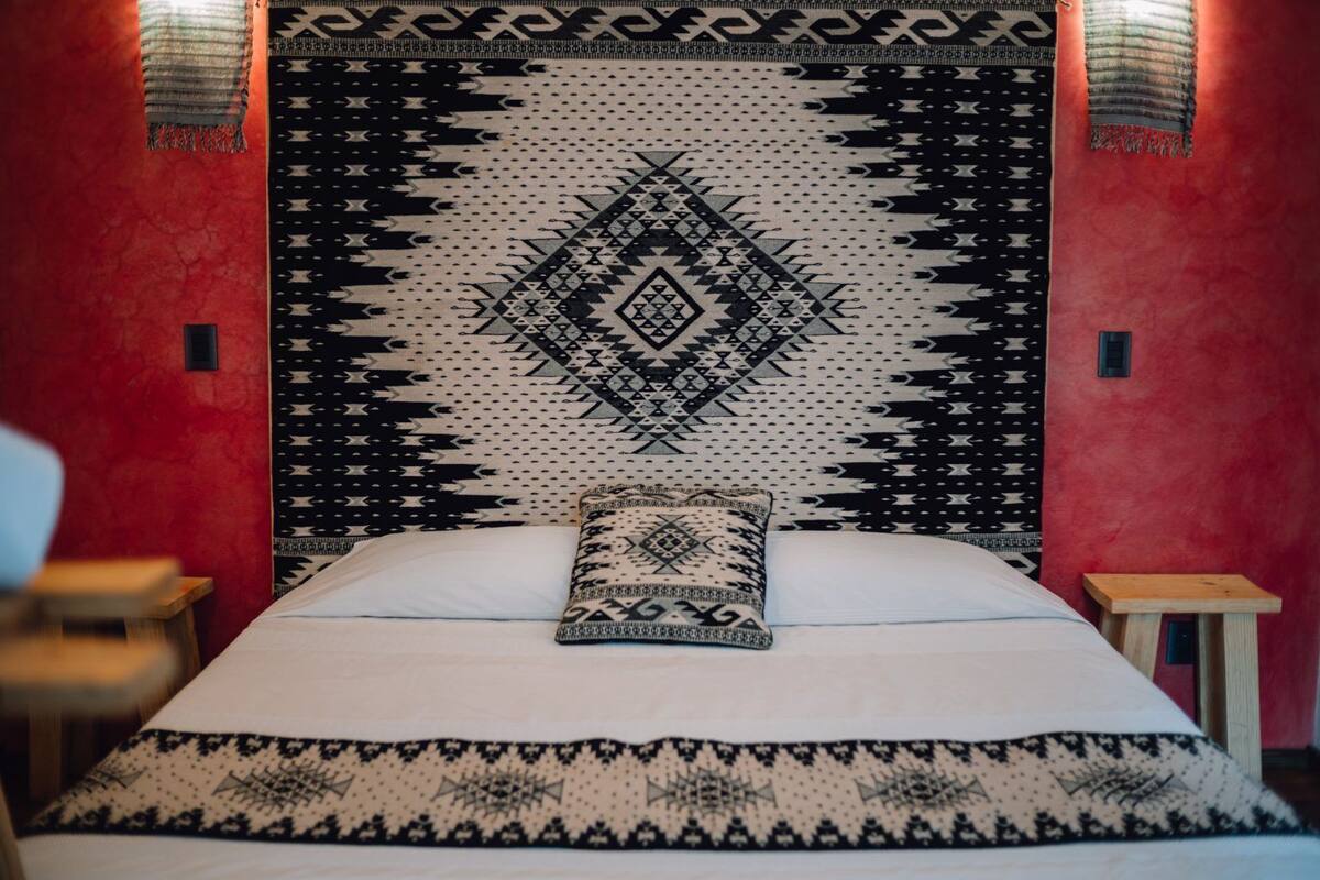 A bed with a patterned headboard and pillows, a wooden nightstand, and a red wall.