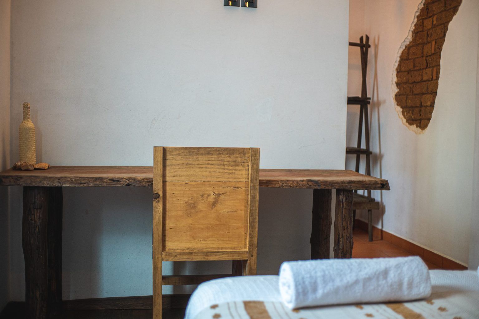 A rustic wooden table with a chair, a towel on the bed, and a brick wall.