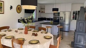 Un coin repas avec une table ronde dressée pour six personnes, entourée de chaises en bois. Un luminaire suspendu noir se trouve au-dessus de la table, et un vase rempli de fleurs est posé sur la table.