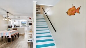 Un escalier aux marches bleues menant à un palier, un coin repas avec une table et des chaises, et une cuisine visible au fond.