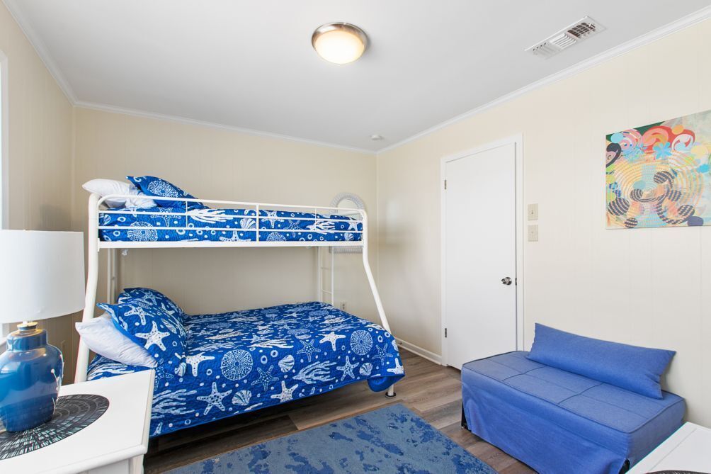 Una cama litera con ropa de cama azul y blanca de temática náutica, una cama individual con ropa de cama a juego, un sofá azul, una lámpara y un cuadro abstracto y colorido en la pared.