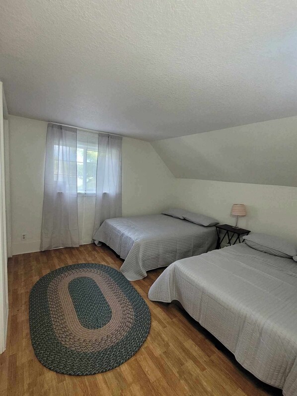 A small, single-room accommodation with two beds, a window with sheer curtains, a small table with a lamp, and a round rug on a wooden floor.