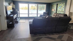 A living room with a dark-colored sofa, a coffee table, and a TV on a stand. There is a large window with a view of a body of water and trees. - Lake Front on Little Clear Lake- Comfort Resort (Prescott)