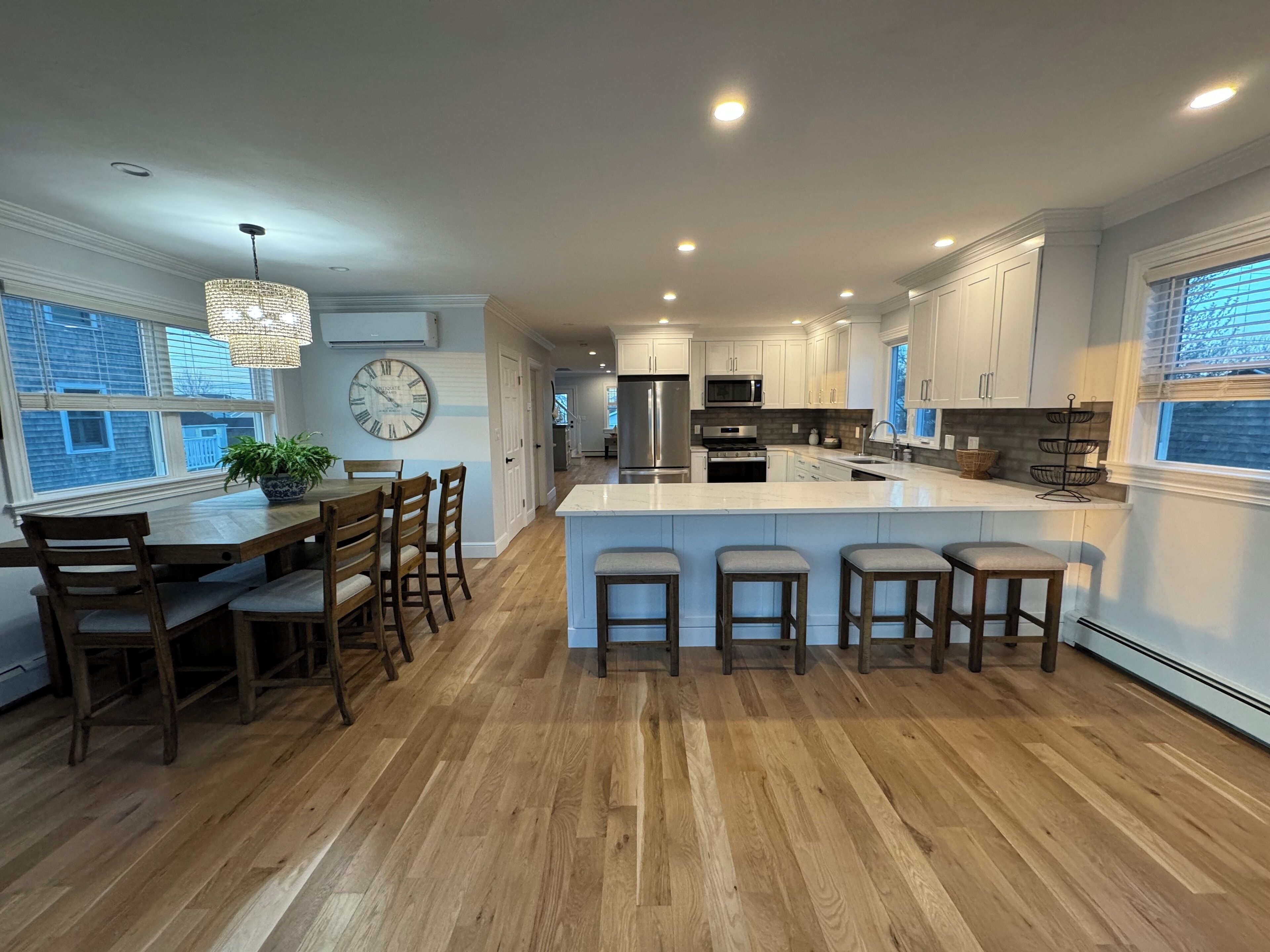 Une cuisine moderne avec un plancher en bois, des armoires blanches, un grand îlot central doté de tabourets, et un coin repas avec des chaises en bois.