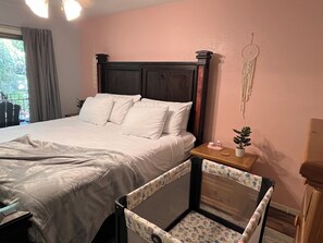 Una cama bien hecha con sábanas blancas, un cabecero de madera, una mesita de noche con una planta pequeña y un adorno, y una ventana con cortinas.
