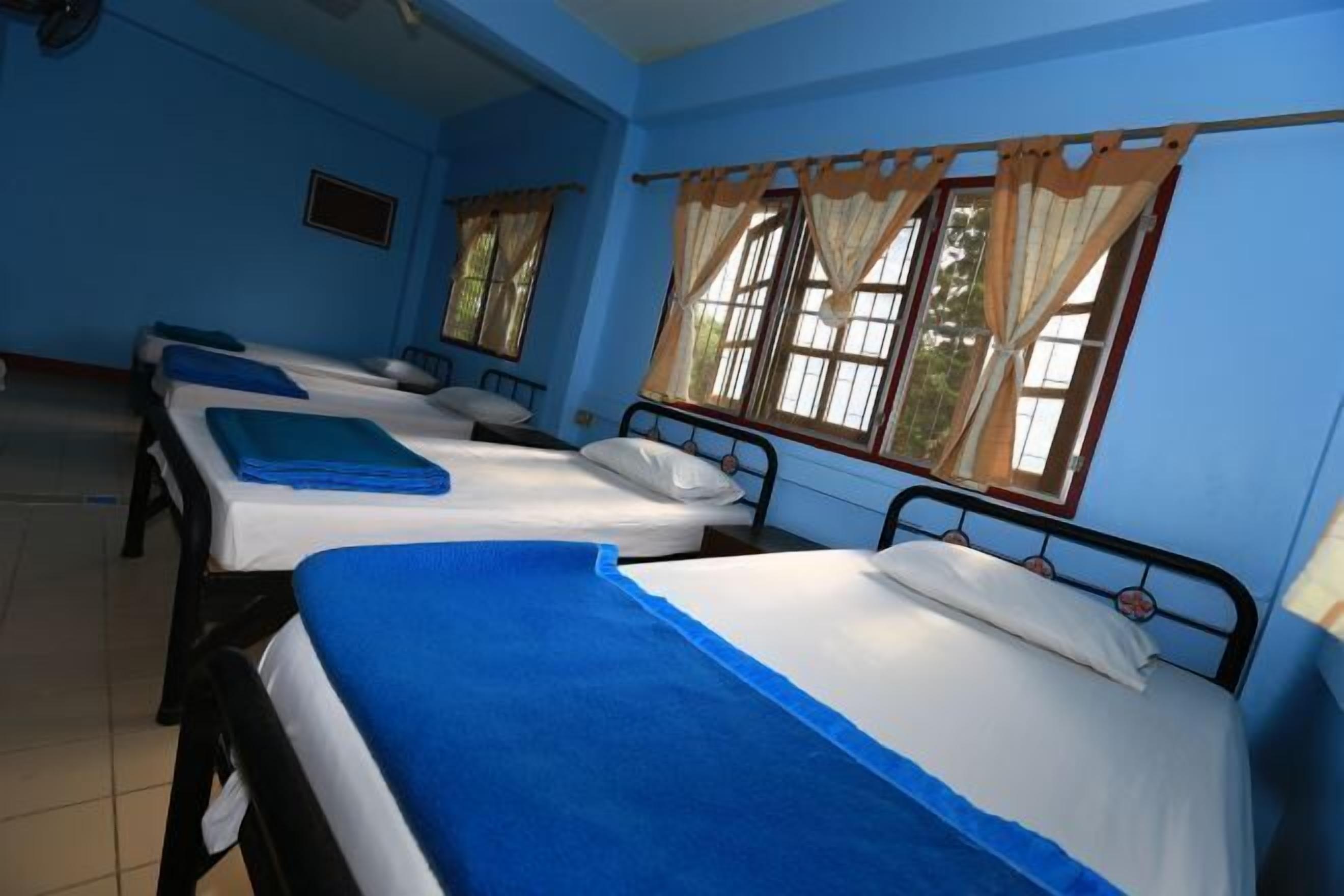 A room with three single beds, each with a blue bedspread and white sheets. There are windows with curtains, and the walls are painted blue.