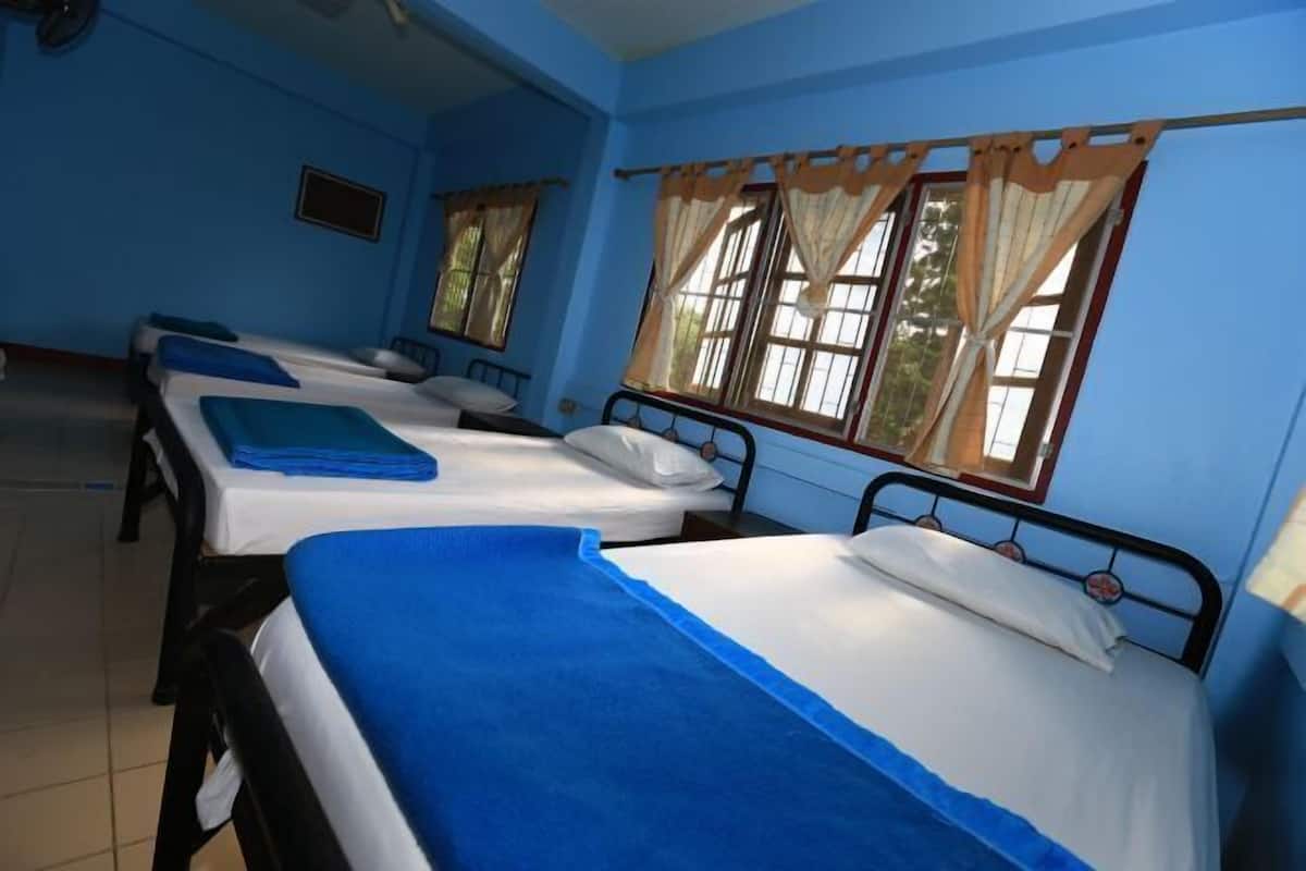 Een kamer met drie eenpersoonsbedden, elk met een blauwe sprei en witte lakens. Er zijn ramen met gordijnen, en de muren zijn blauw geschilderd.