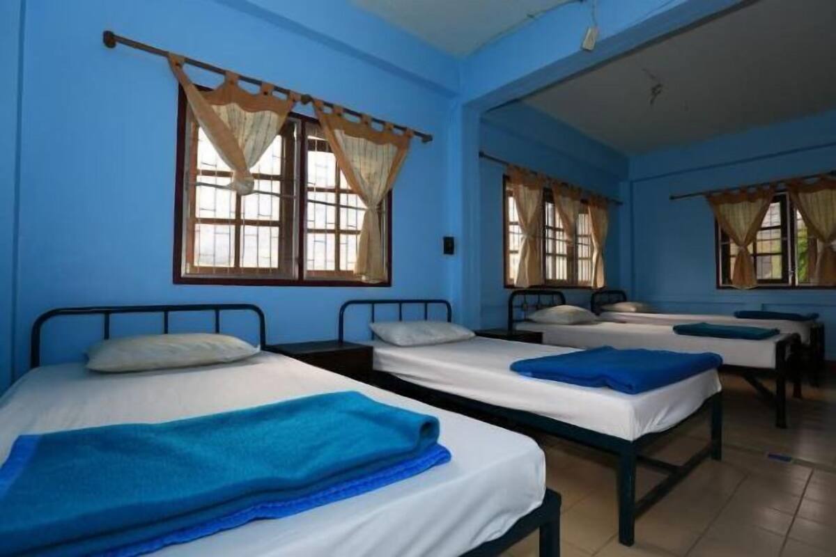 Een kamer met vier eenpersoonsbedden, elk met een blauwe deken, en een raam met gordijn.