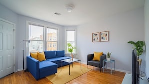 A modern living room with a blue sectional sofa, a small coffee table, and a grey armchair. There are two windows providing natural light, a flat-screen TV, and a potted plant. - 2F - ❤️ Lowest Rates! Fun & Stylish 3BR in Logan Square (Chicago)