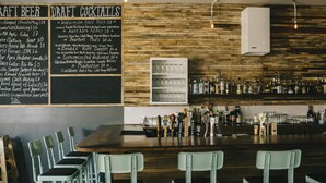 A bar area with a wooden wall, a chalkboard menu listing draft beers and cocktails, and a bar stocked with bottles and glasses. - 2F - ❤️ Lowest Rates! Fun & Stylish 3BR in Logan Square (Chicago)