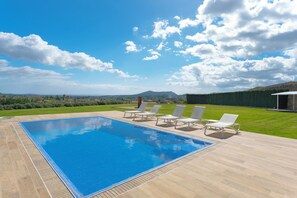 A rectangular swimming pool with a tiled deck, surrounded by white lounge chairs, set against a backdrop of a clear sky and distant mountains. - Casa con Piscina en Selva (Selva)