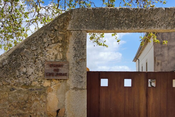 A stone archway with a wooden sign, a metal gate, and a building in the background. - Casa con Piscina en Selva (Selva)