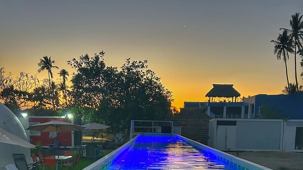 An outdoor swimming pool with a water slide, surrounded by palm trees and a sunset in the background.