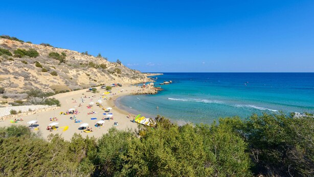 A beach with clear blue waters, sandy shore, and rocky hills in the background. - Villa Amesta - Private Pool Retreat (Protaras)