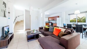 A modern living room with a sectional sofa, a coffee table, and a TV on a stand. There is a staircase leading to an upper level, a kitchen area, and a dining area with a table and chairs. - Villa Amesta - Private Pool Retreat (Protaras)