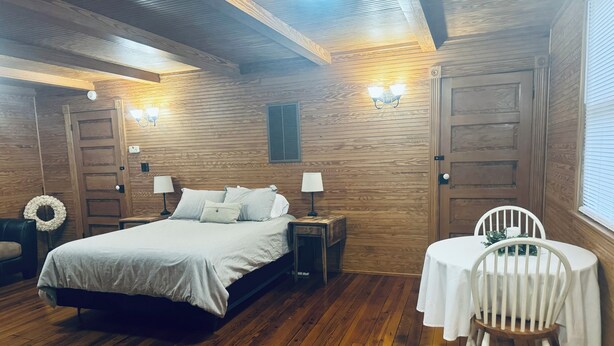 A wooden-paneled room with a bed, bedside lamps, a round table with a chair, and a door with a wreath. - Cozy studio rental situated between Bedford and Lynchburg Virginia (Forest)