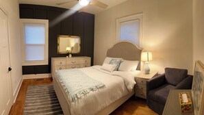 Une chambre à coucher comprenant un lit, des tables de chevet, une chaise, une commode, un miroir et une fenêtre avec des stores.