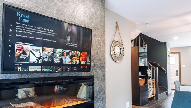 Ein modernes Wohnzimmer mit Kamin, einem an der Wand befestigten Fernseher, der einen Film zeigt, und einer Treppe, die zu einer anderen Ebene führt.
