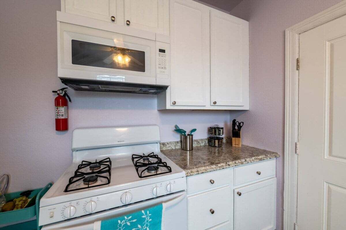 Una cocina compacta con armarios blancos, estufa a gas, microondas y extintor.