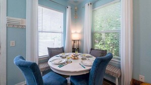 A dining area with a round table set for four, blue upholstered chairs, white curtains, and a lamp between the windows.